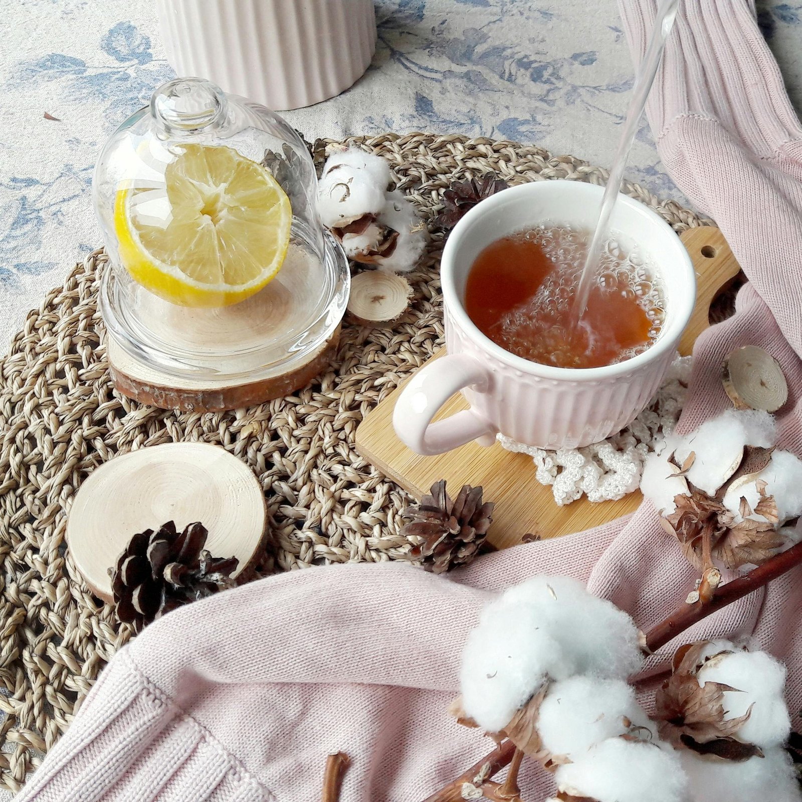A warm tea being poured into a white mug on a woven placemat, accompanied by lemons, pine cones, and cotton for a cozy winter feel.